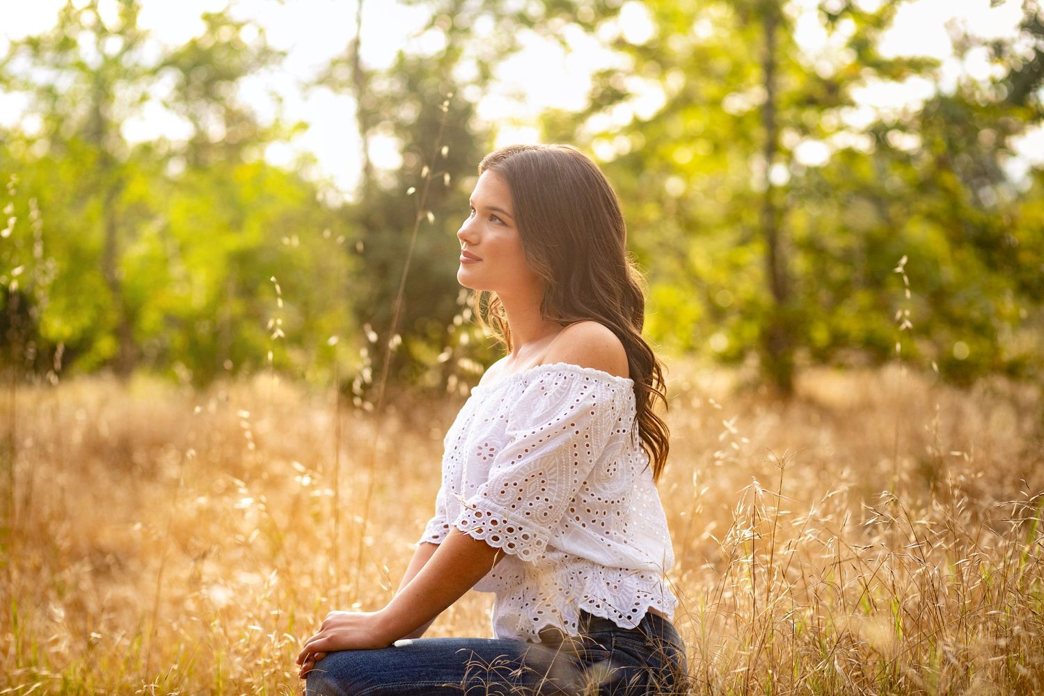 Young woman in white off-shoulder top and jeans sitting in golden meadow with green trees in sunlit background.