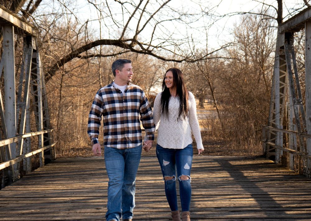 Couple holding hands walking on a rustic wooden bridge surrounded by bare autumn trees in warm golden light.