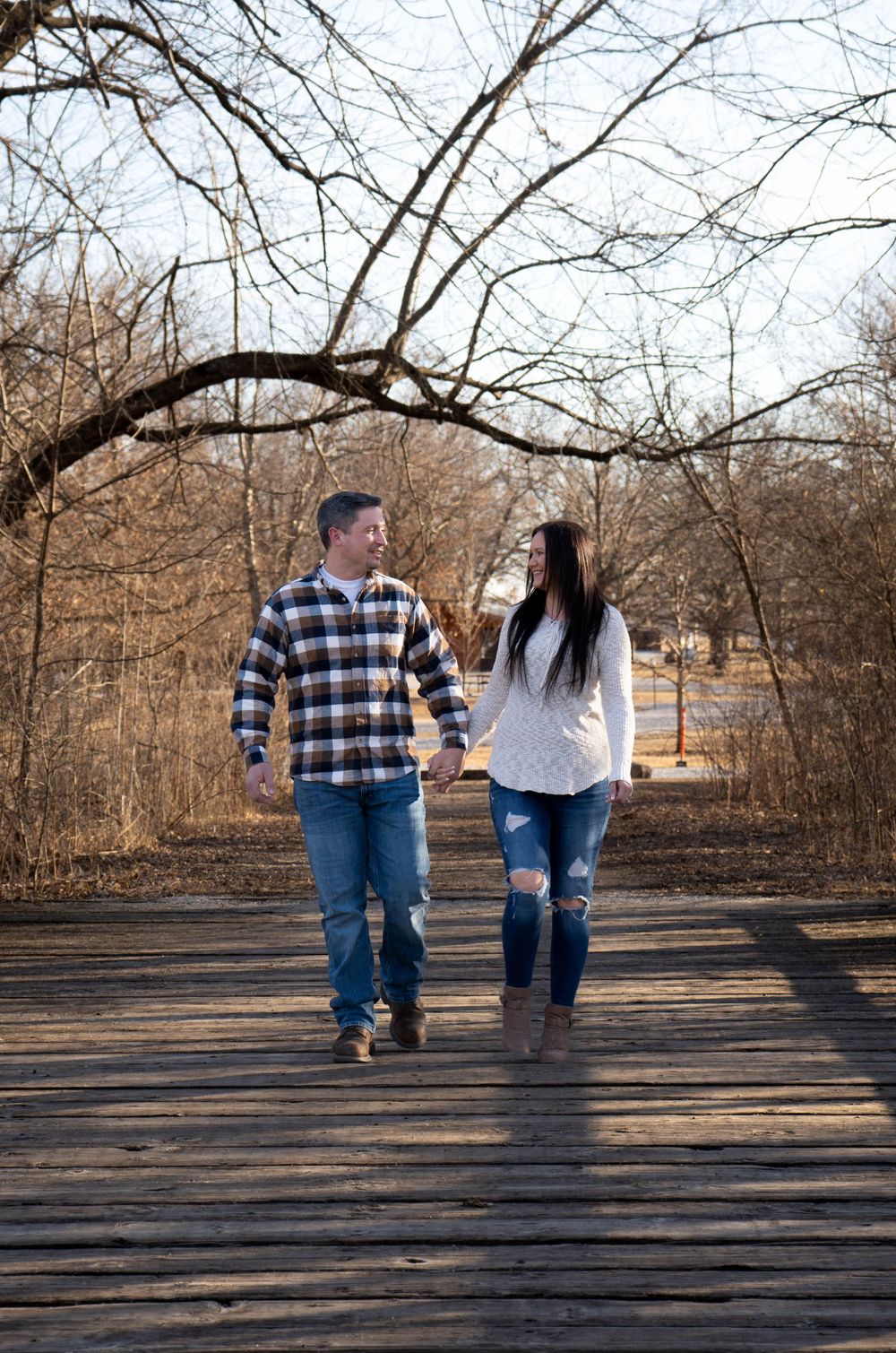 Happy couple holding hands strolling on a rustic wooden boardwalk surrounded by bare trees.