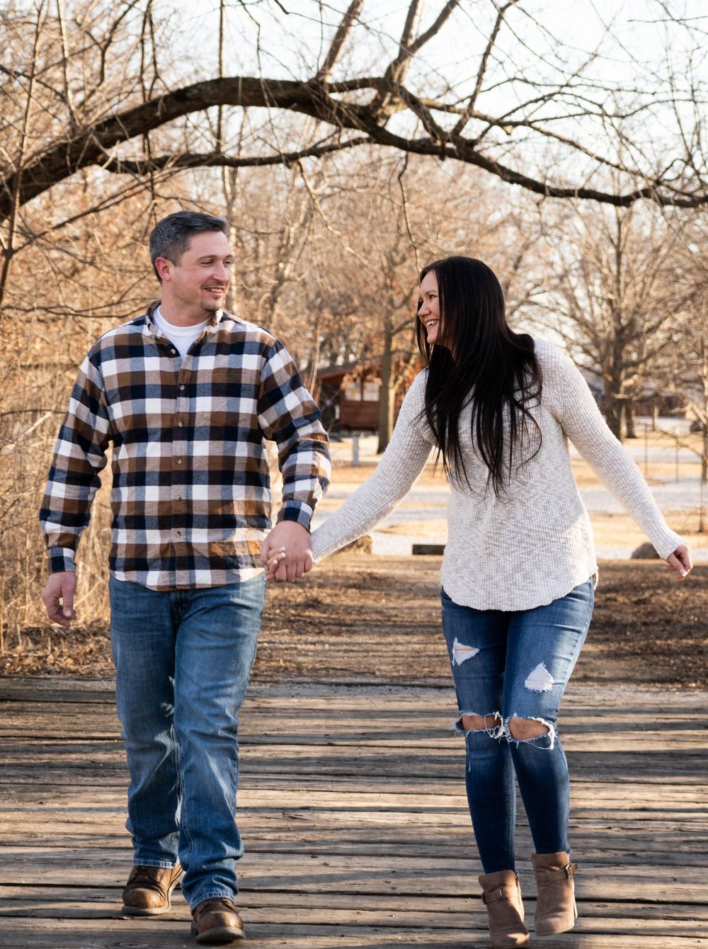 Couple holding hands and smiling while walking on a wooden path outdoors in autumn.