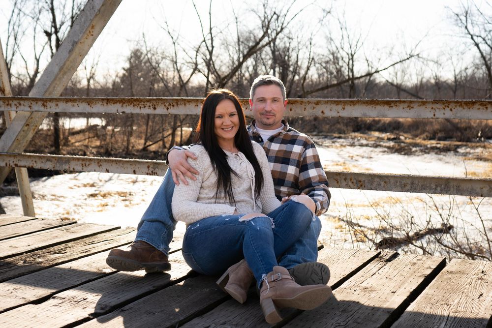 Couple sitting on wooden bridge smiling, woman in cream sweater and jeans, man in plaid shirt, winter outdoor setting.