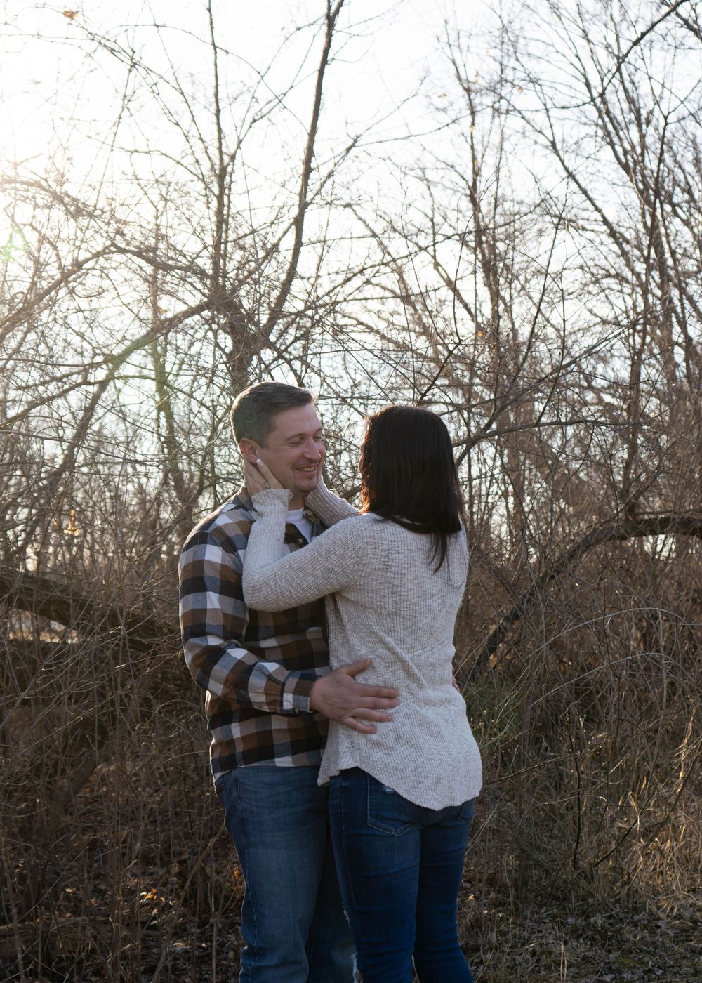Couple embracing outdoors among bare winter trees, wearing casual flannel and knit sweater, in a romantic engagement photo.