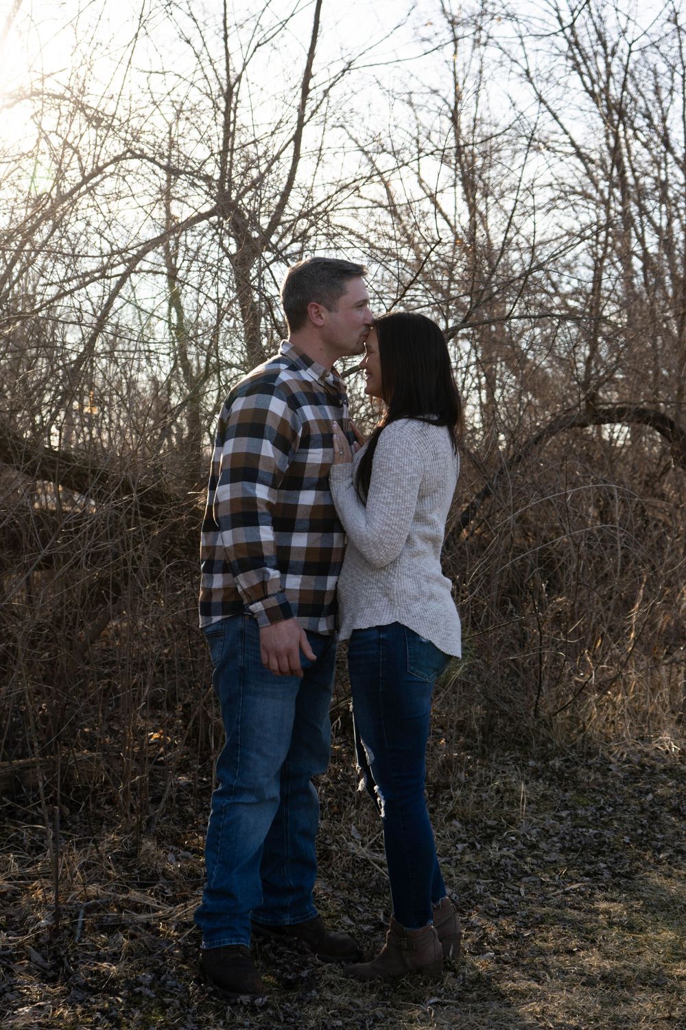 Couple sharing a romantic moment outdoors in winter, surrounded by bare trees, wearing casual flannel and sweater.