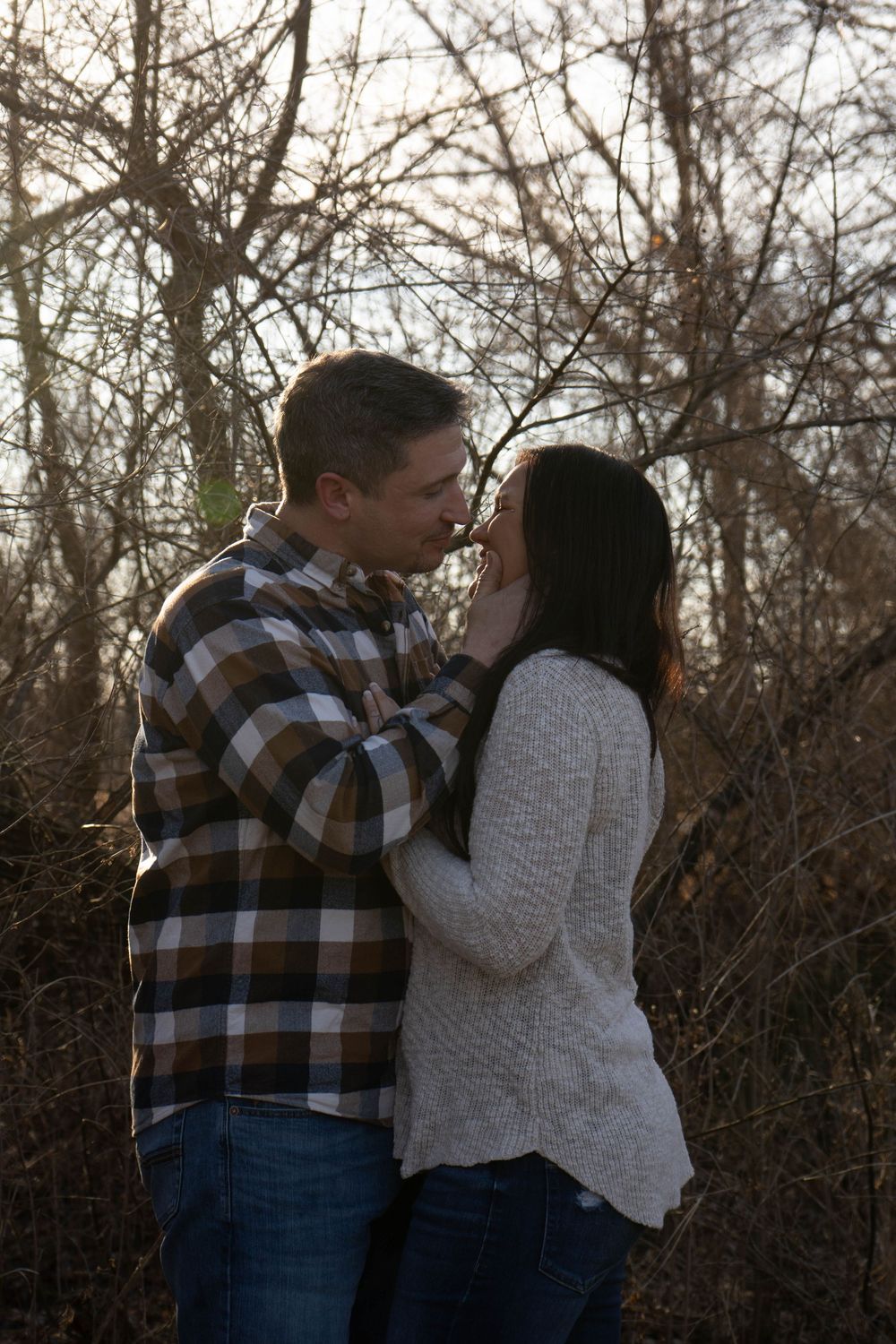 A couple sharing a tender kiss surrounded by bare winter trees in a romantic outdoor setting.