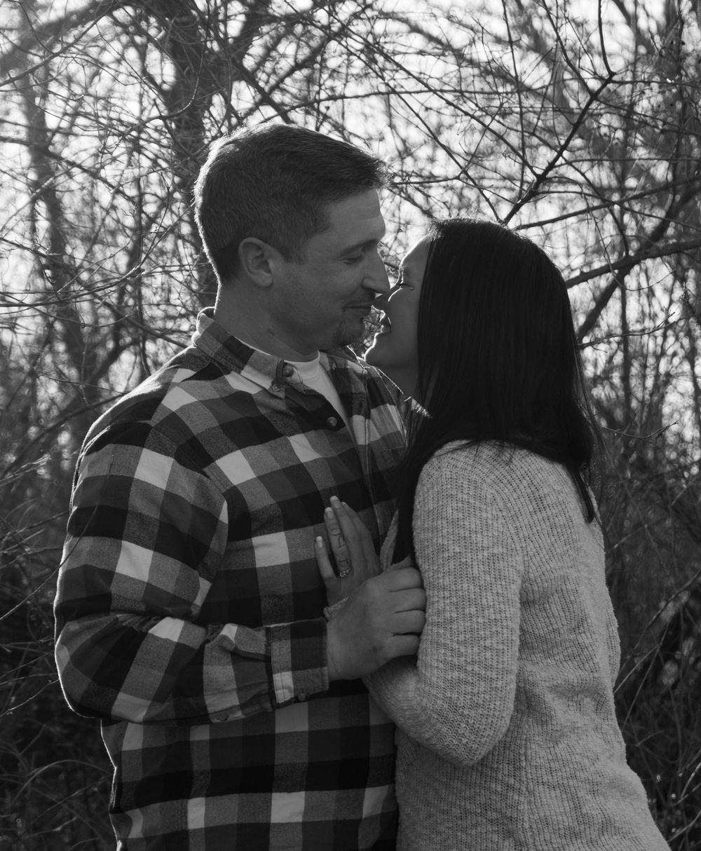 Black and white photo of a smiling couple embracing outdoors among bare trees in a romantic moment.