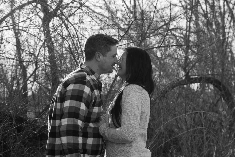 Black and white photo of a romantic couple sharing a tender moment outdoors among bare winter trees.