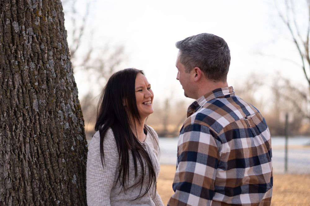 Couple smiling at each other beside a large tree in a park during autumn, woman with long dark hair, man in plaid shirt.