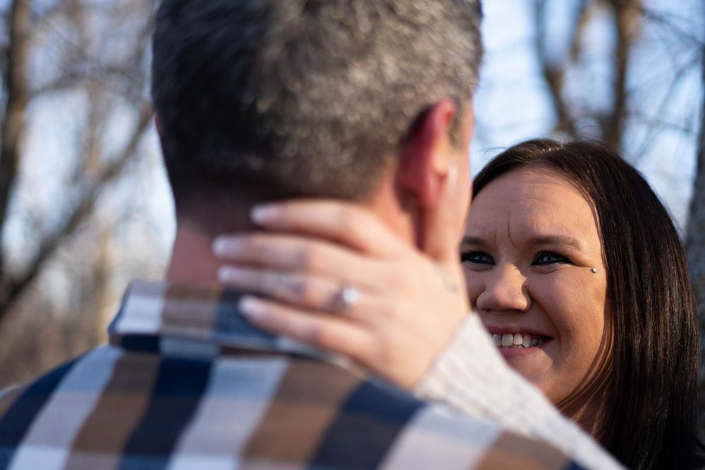 A smiling woman with an engagement ring embraces a man outdoors among bare trees in soft natural light.