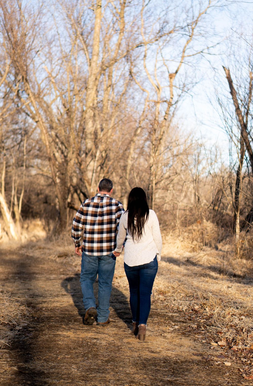 A couple holding hands walking down a rustic dirt path lined with bare autumn trees.