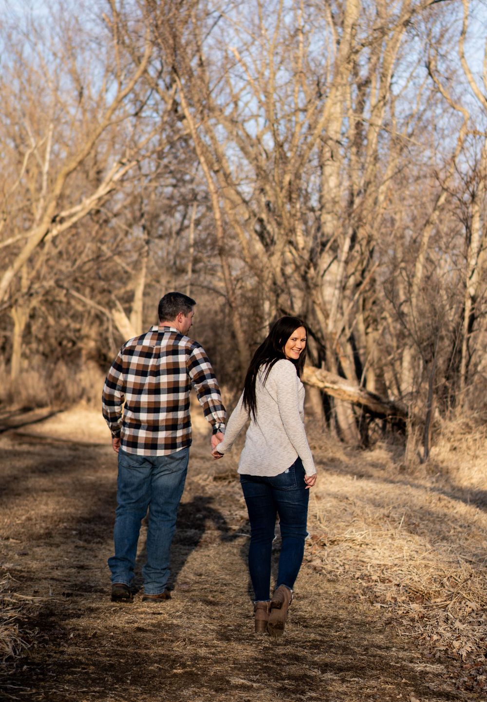 Couple walking hand-in-hand on autumn trail through bare trees, woman glancing back smiling, casual outdoor engagement photo.