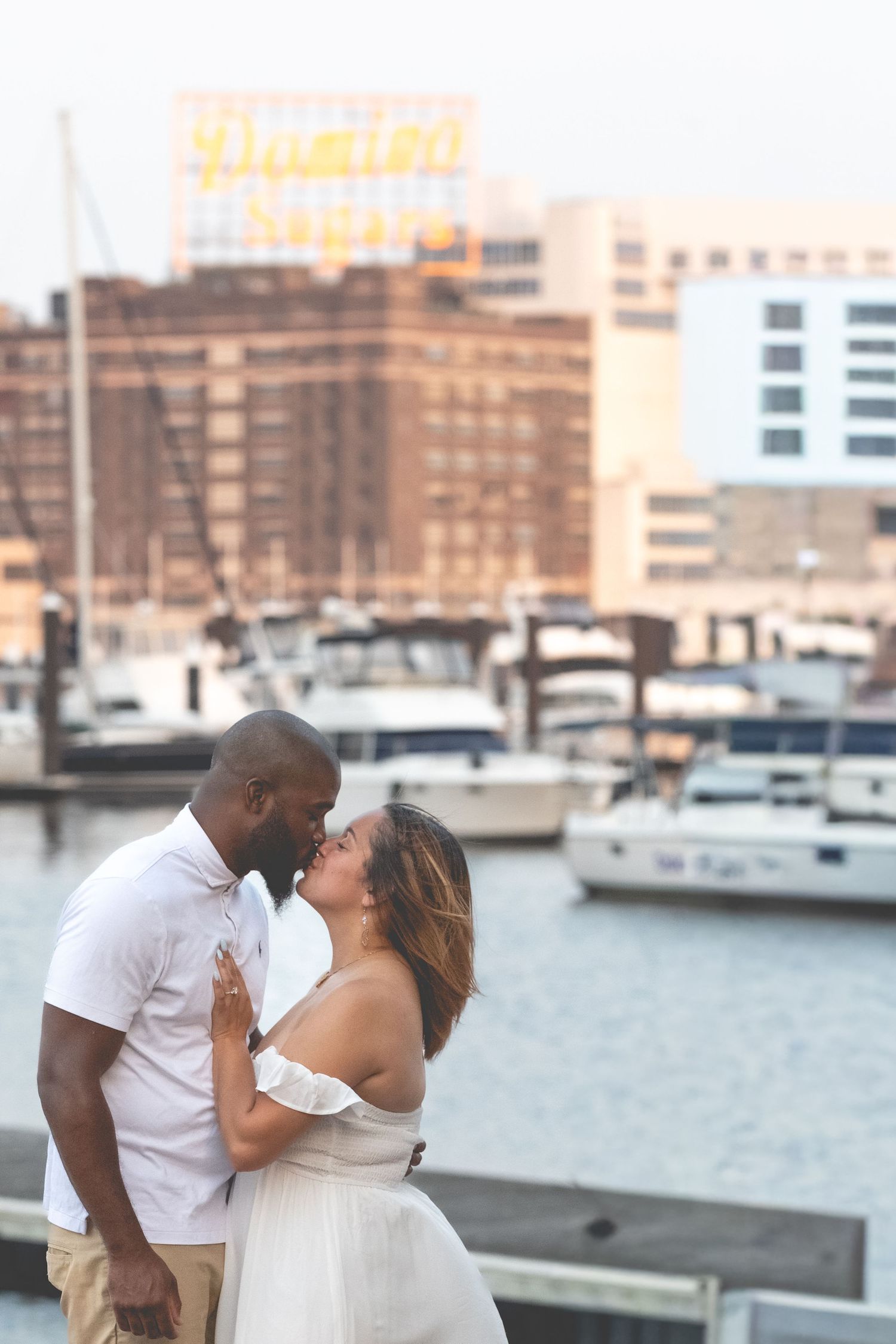 Interracial couple sharing a kiss at a harbor with boats and city skyline behind them.