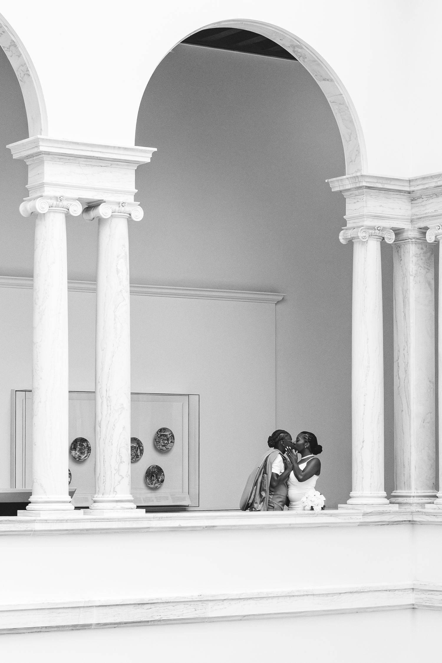 Black and white photo of a couple sharing a tender moment beneath classical arched columns.