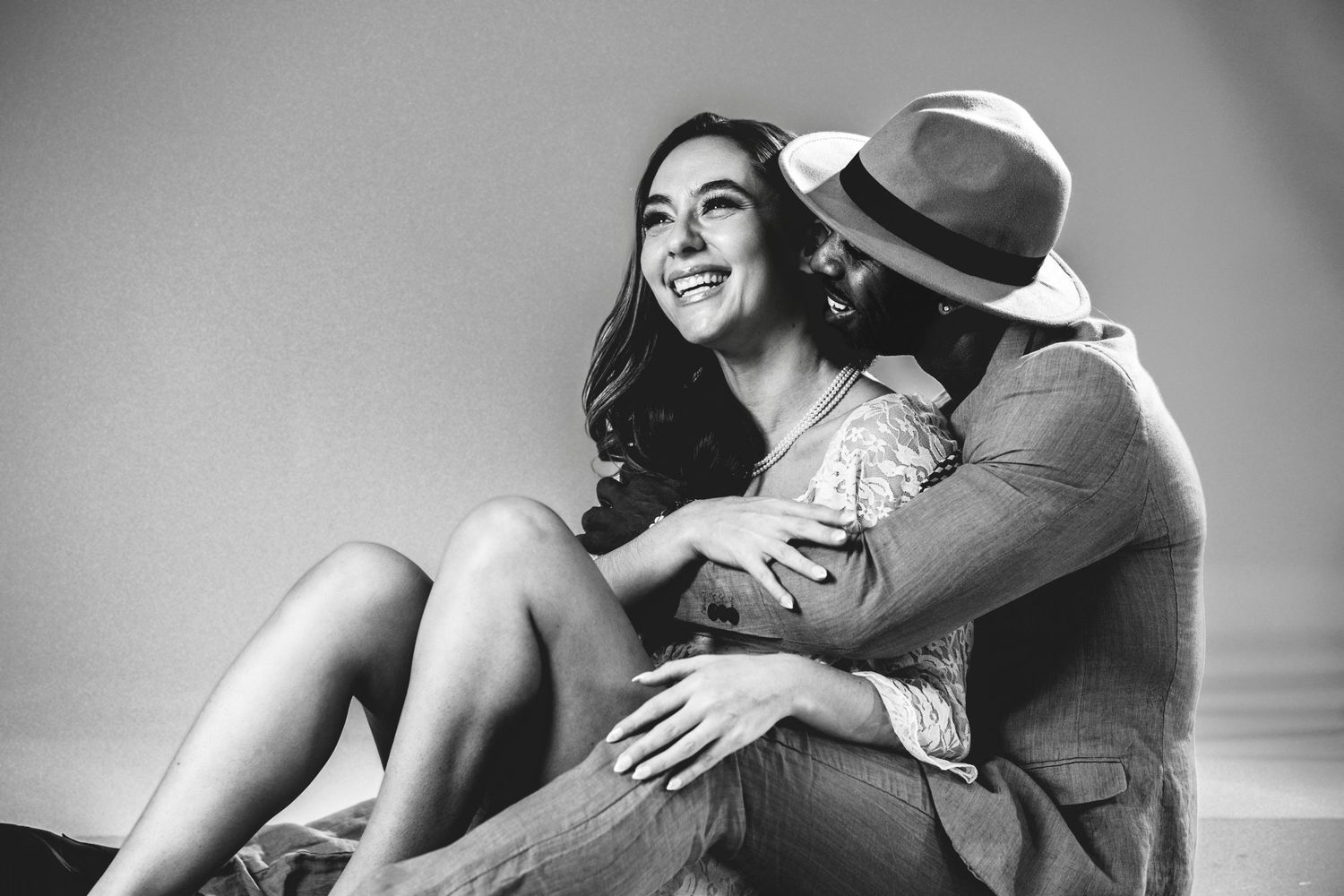 Black and white photo of a happy couple sitting together, woman laughing joyfully as man in hat embraces her tenderly.