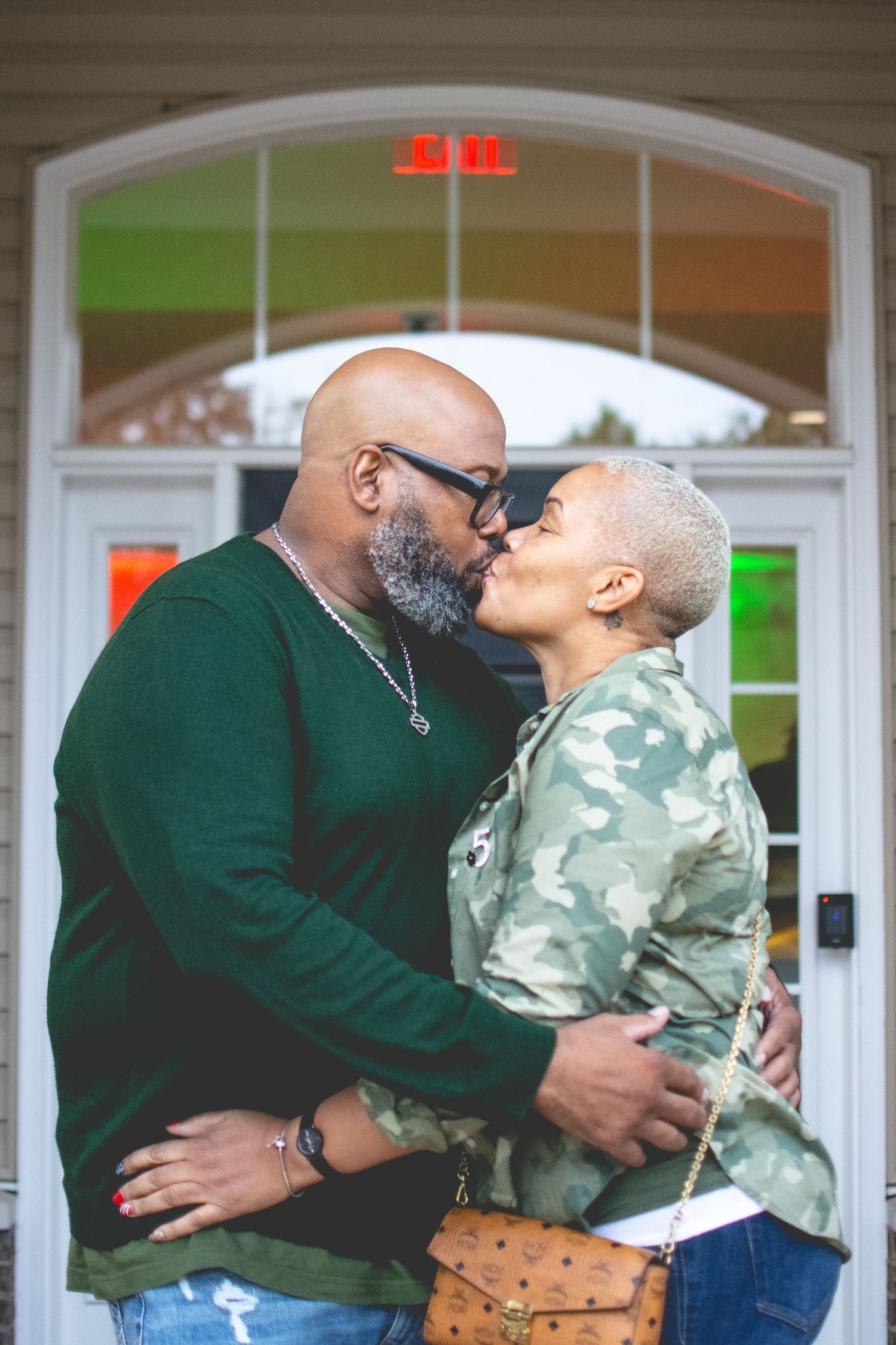 A couple shares a romantic kiss outside a building, man in green sweater and woman in camo jacket with short blonde hair.
