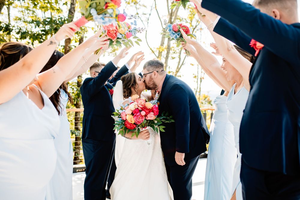 Bride and groom share a kiss under an arch of raised flowers held by wedding party members outdoors.