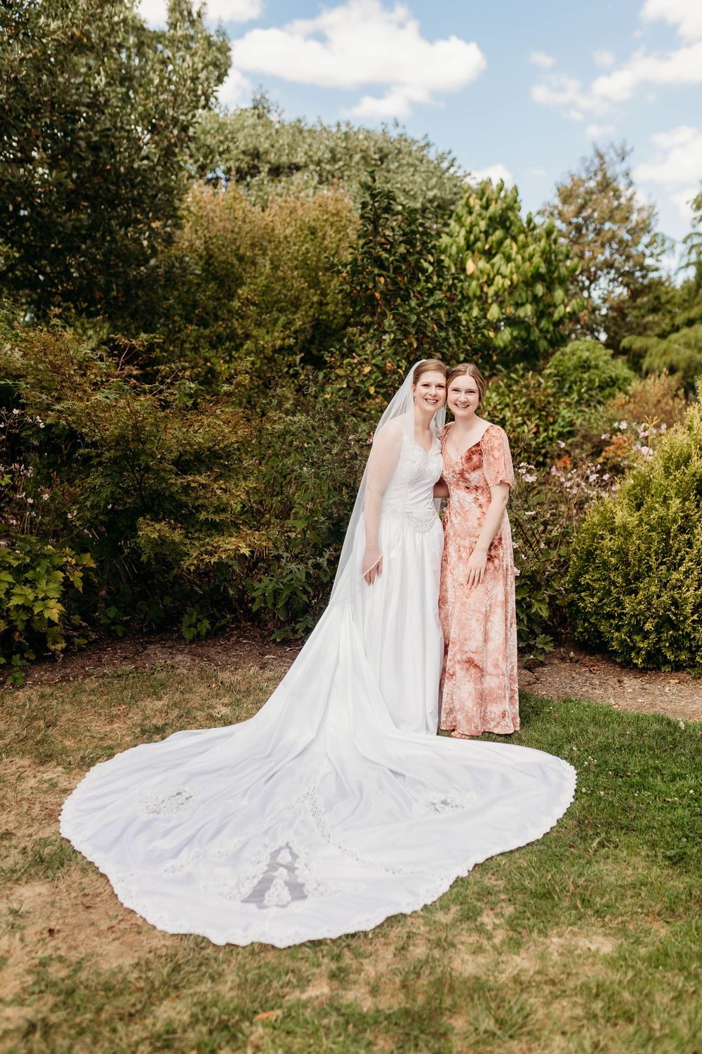 Bride in white gown with long veil poses with bridesmaid in floral dress in lush garden setting.