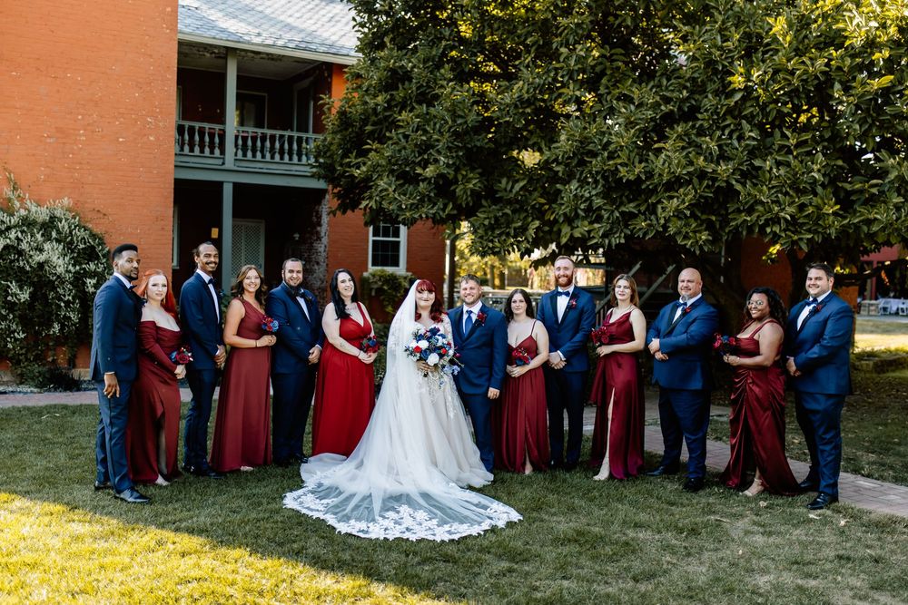 Wedding party in burgundy and navy attire posing outdoors on green lawn beside brick building.