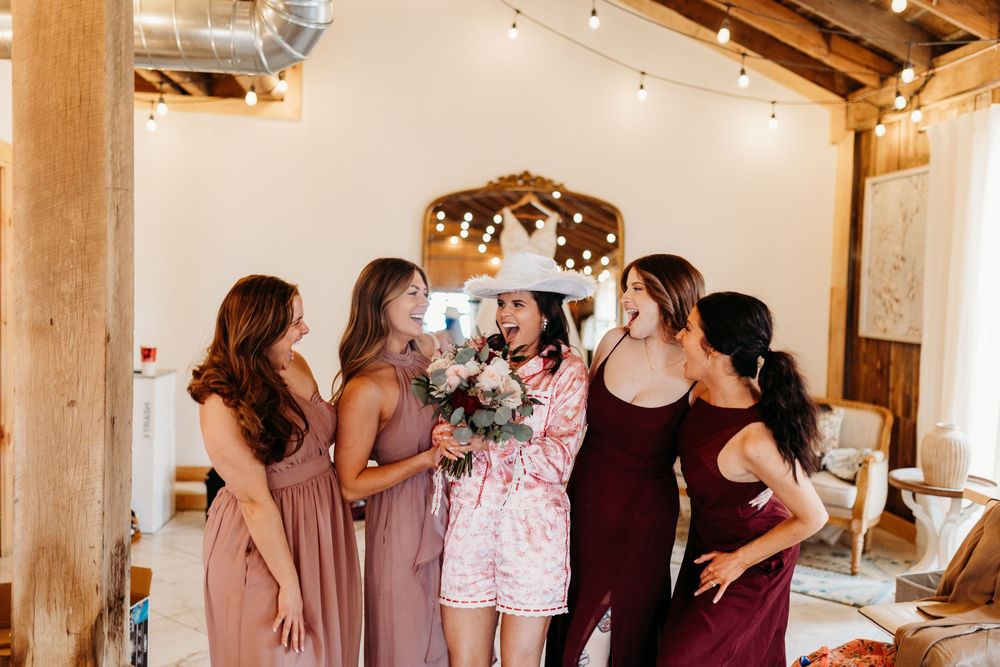 Bride in floral pajamas laughs with bridesmaids in mauve and burgundy dresses inside rustic venue.