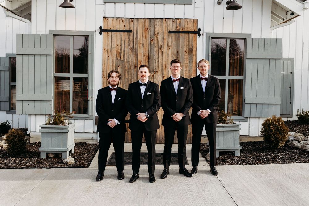 Four groomsmen in black tuxedos with bow ties posing in front of a rustic white barn with wooden doors.