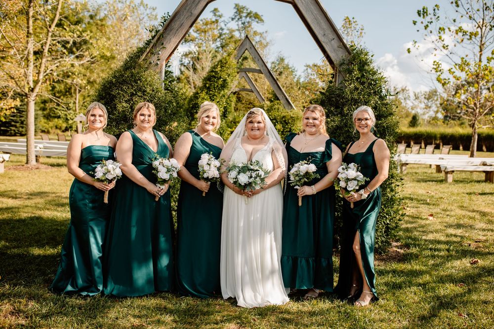 Bride in white gown poses with five bridesmaids in emerald green dresses under a wooden arch outdoors.