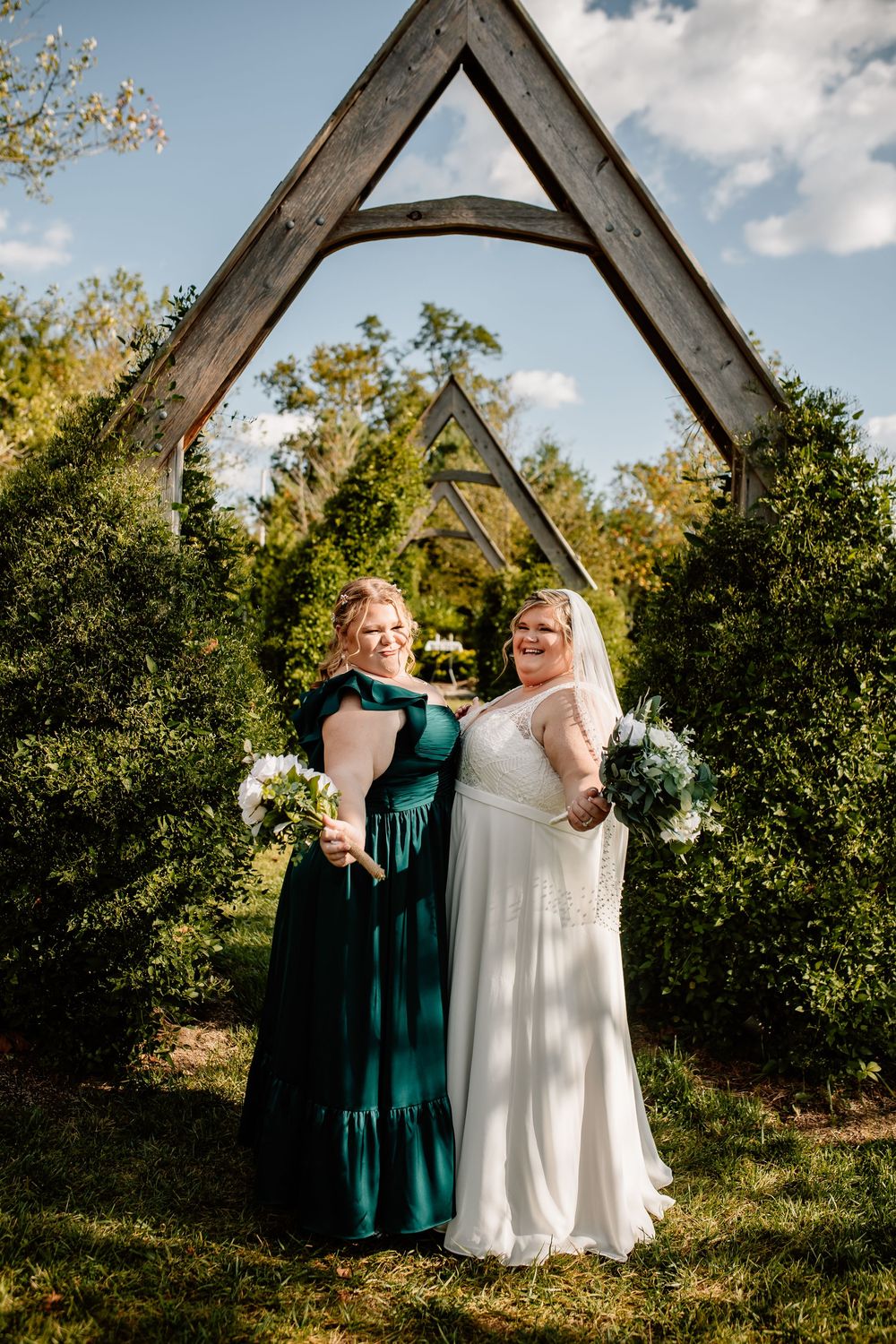 Bride in white lace gown poses with bridesmaid in teal dress under wooden arch outdoors.
