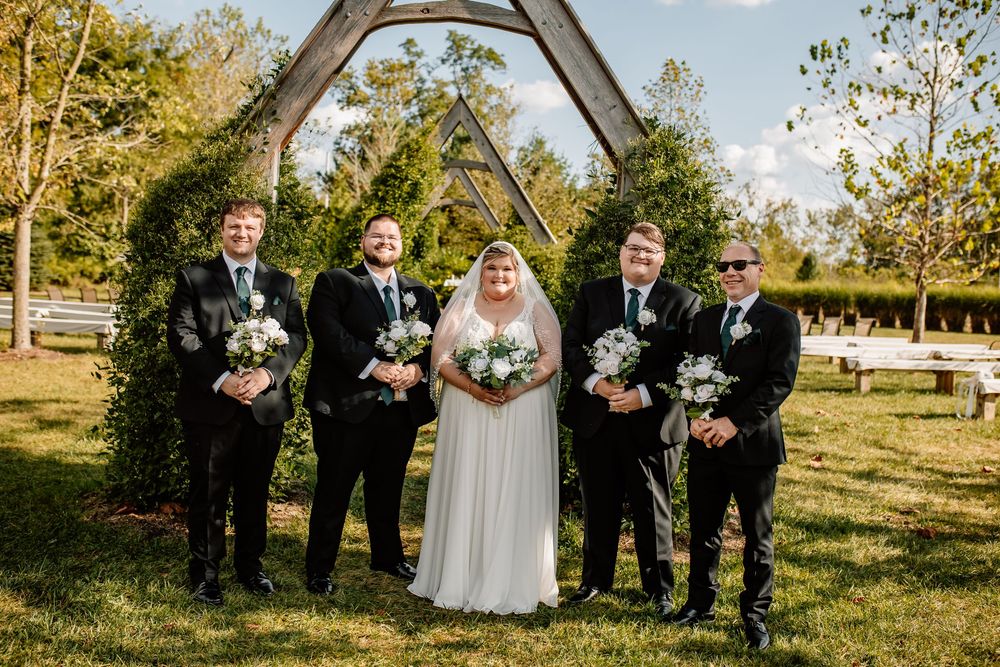 Bride in white gown poses with groomsmen in black suits holding bouquets under wooden arch on green lawn.