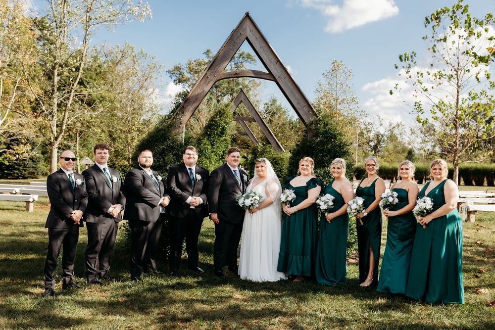 Wedding party in teal and black attire standing before a wooden arch outdoors on a sunny day.