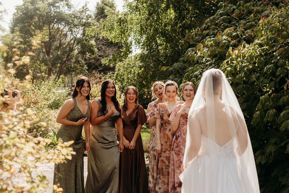 Bridesmaids in mixed earth-tone dresses react with joy as bride in white gown and veil approaches in a garden setting.