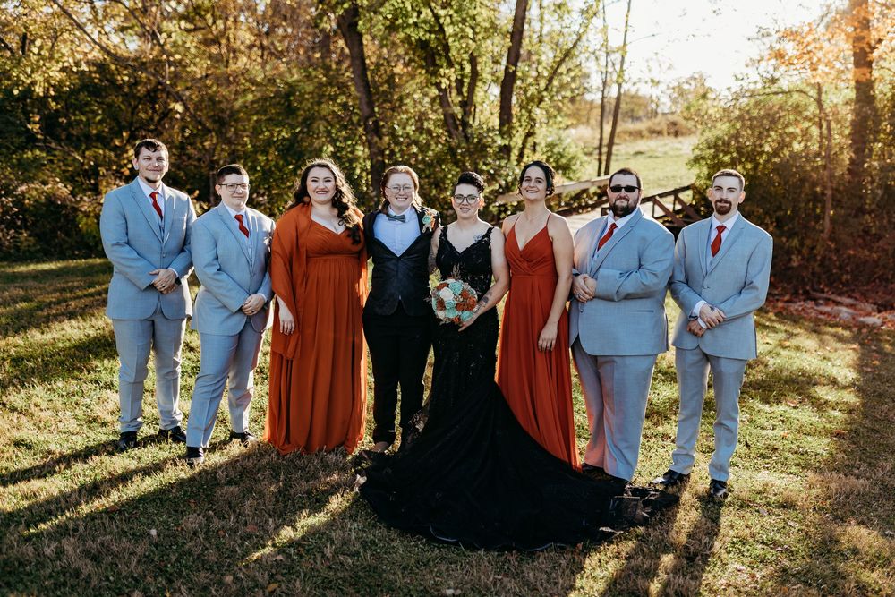 Wedding party poses outdoors in fall, groomsmen in gray suits, bridesmaids in rust dresses, bride in black gown.