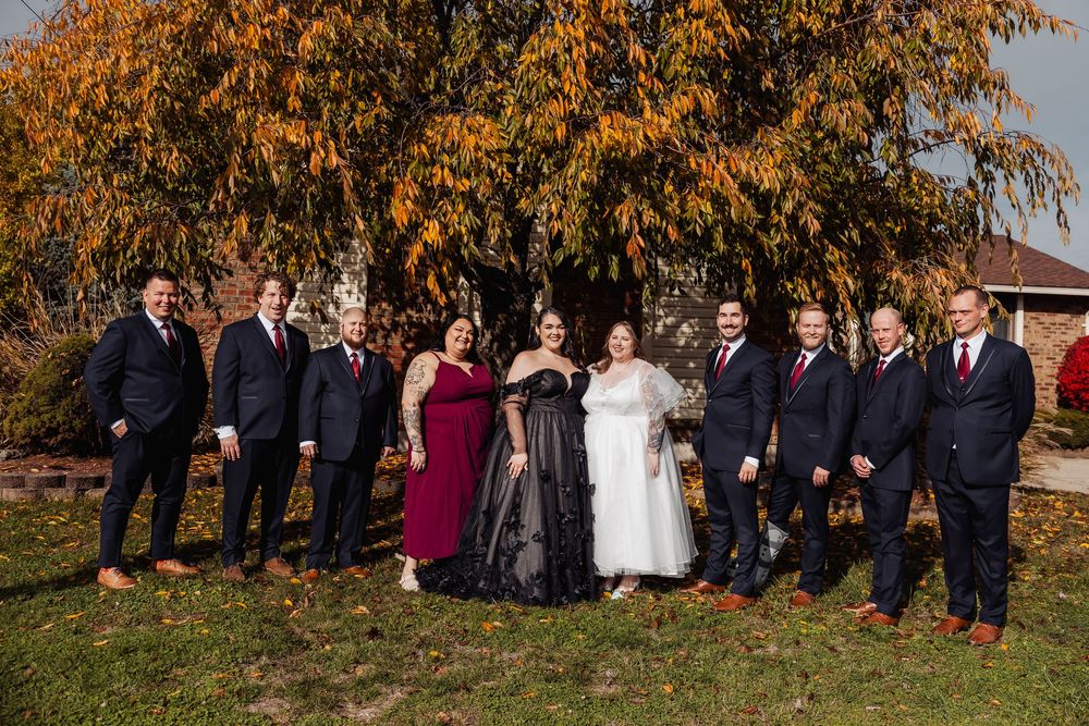 Wedding party poses outdoors in fall foliage, wearing navy suits and burgundy dresses with bride and groom centered.