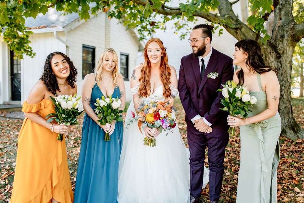 Bride in white gown with groom in burgundy suit and three bridesmaids in colorful dresses holding bouquets outdoors.