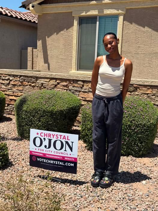 Person standing next to a Chrystal O'Jon for City Council campaign yard sign in front of a house.