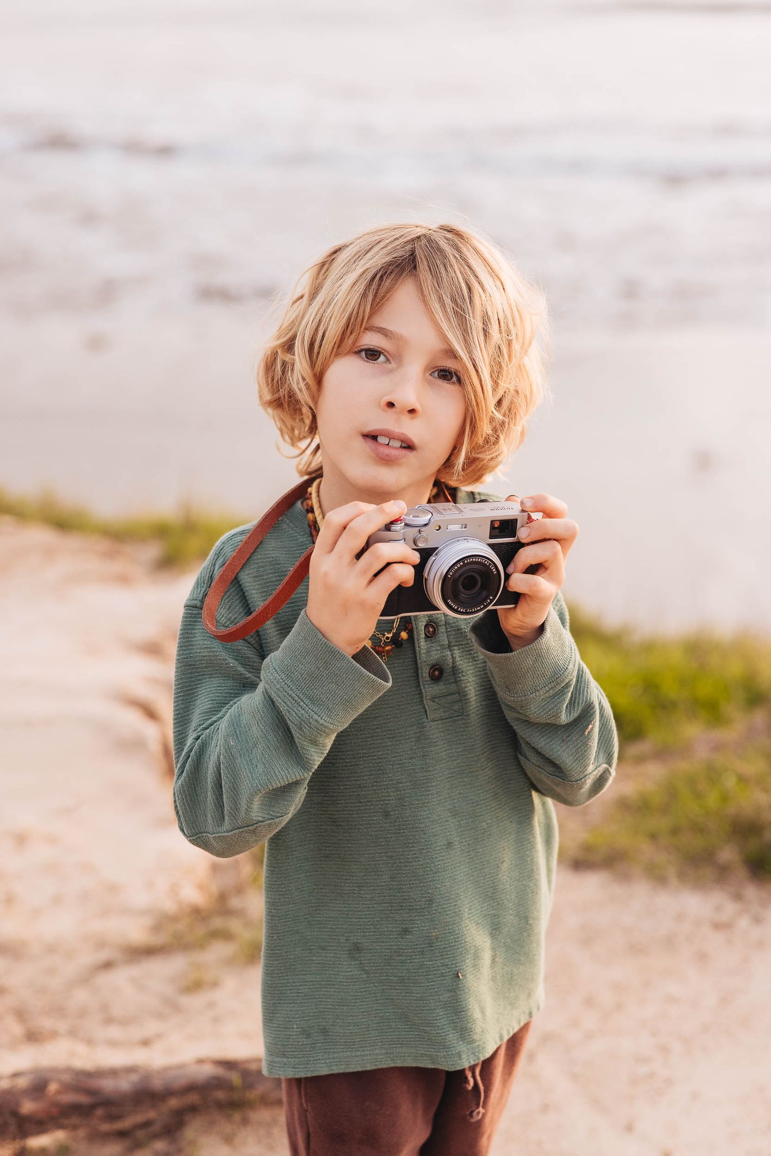 A young boy with blonde hair holds a vintage film camera outdoors near a beach, wearing a green long-sleeve shirt.