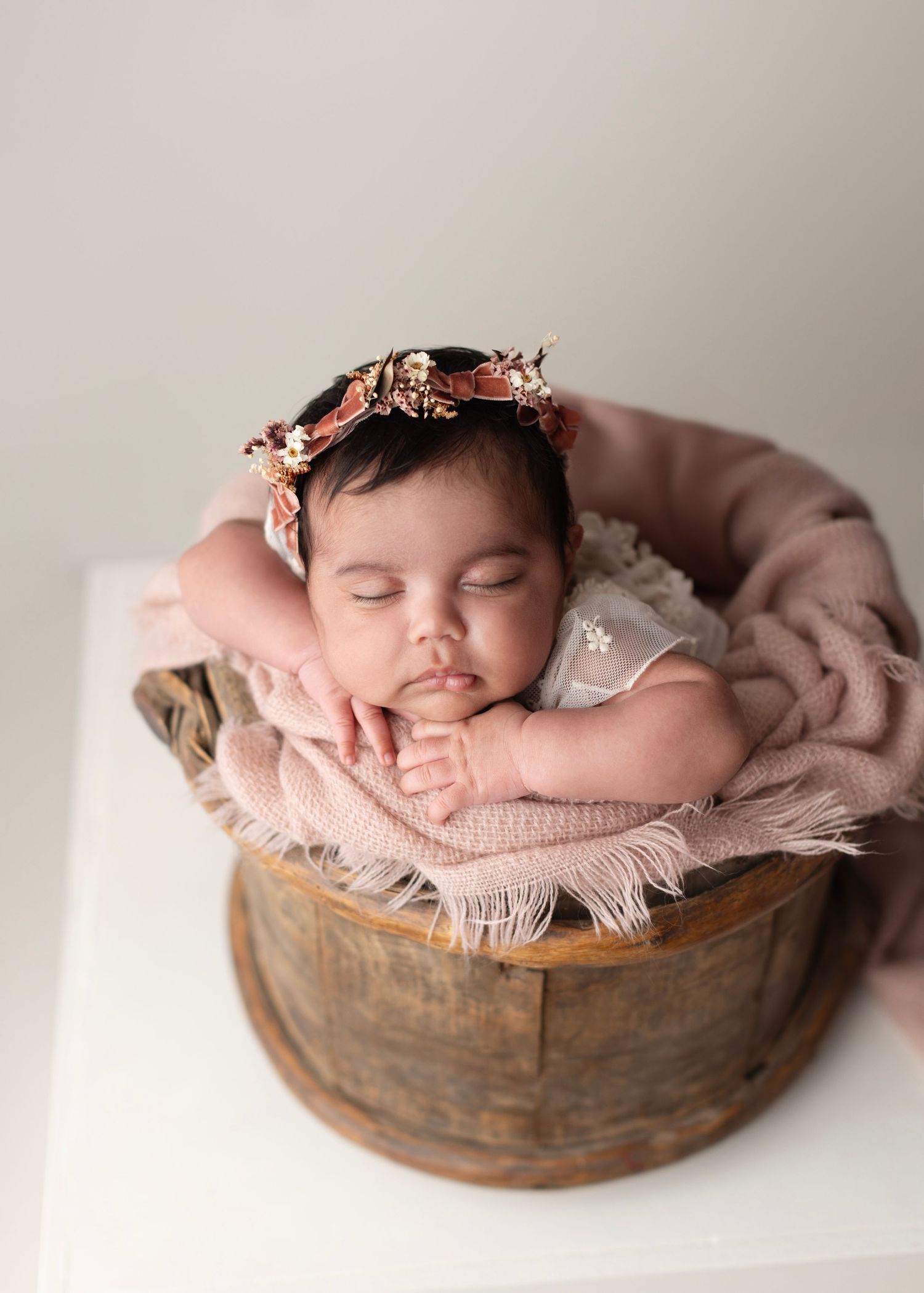 Sleeping newborn baby in wooden bucket with pink blanket and floral headband, peaceful studio portrait.