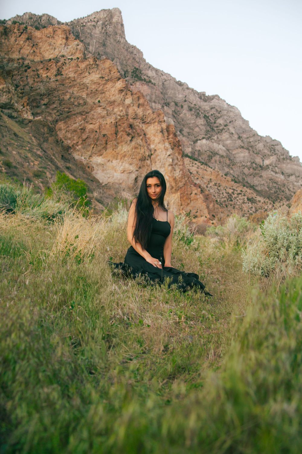 Woman in black dress sitting gracefully in tall grass with dramatic rocky mountain behind her.