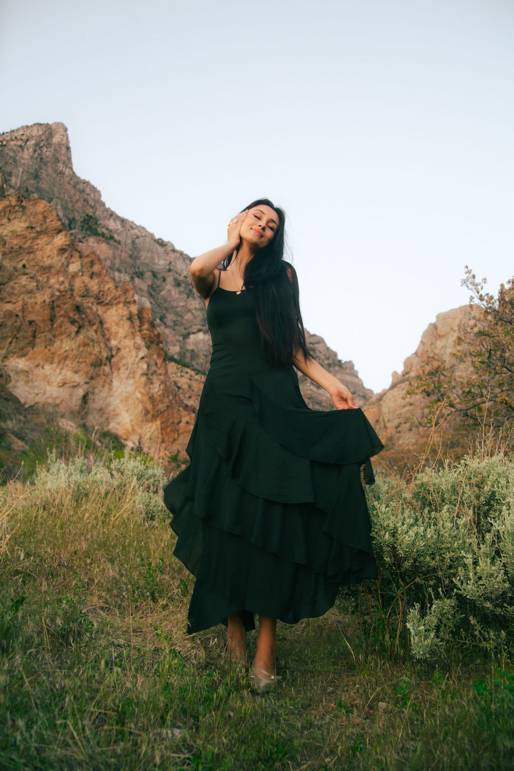 Woman in layered black ruffled dress posing with eyes closed near rocky mountain landscape.