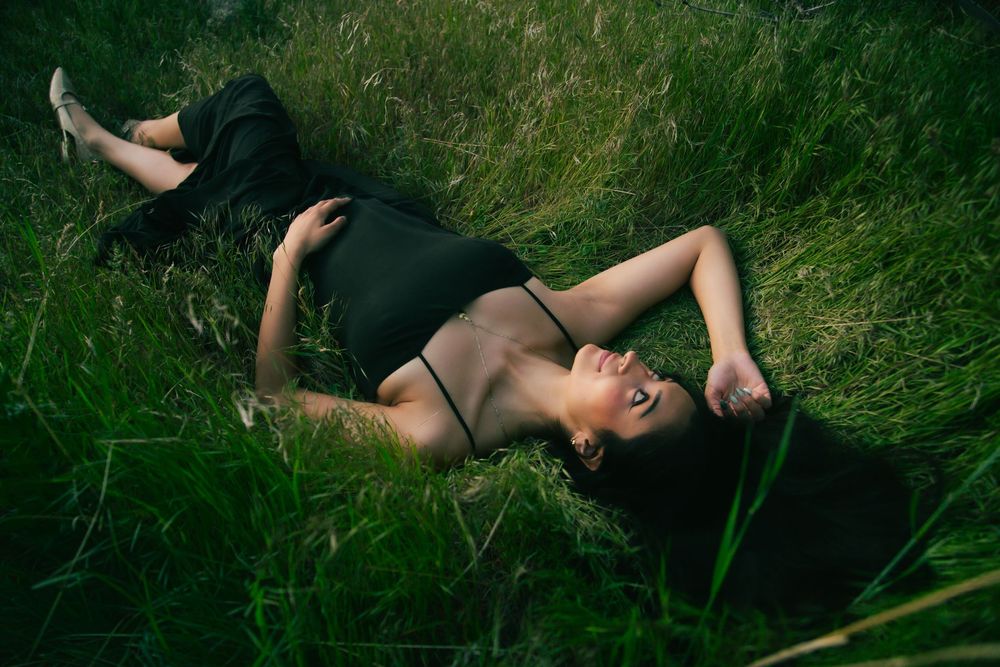 Woman in black dress lying peacefully in lush green grass with eyes closed.