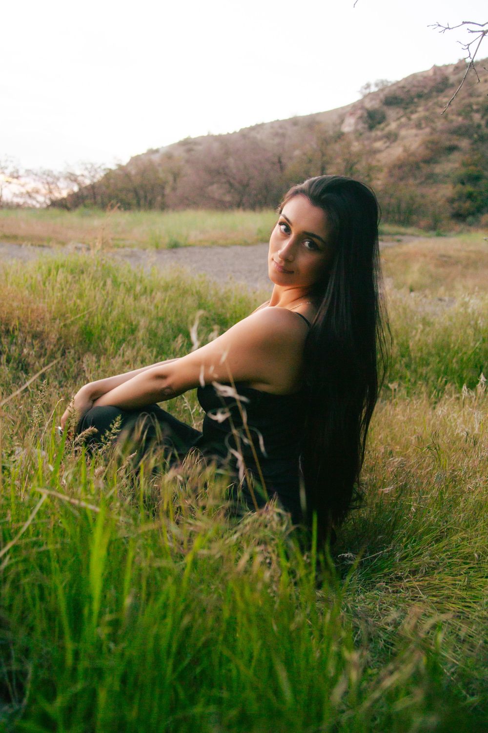 Young woman with long black hair in black dress sits in green grass with hills in background.