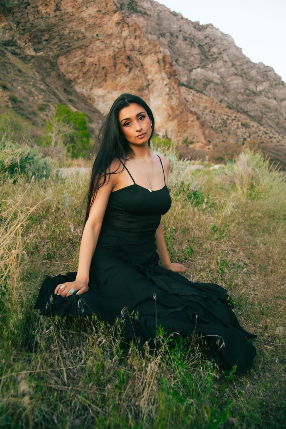Woman in elegant black spaghetti strap dress sits in field with rocky mountain backdrop.