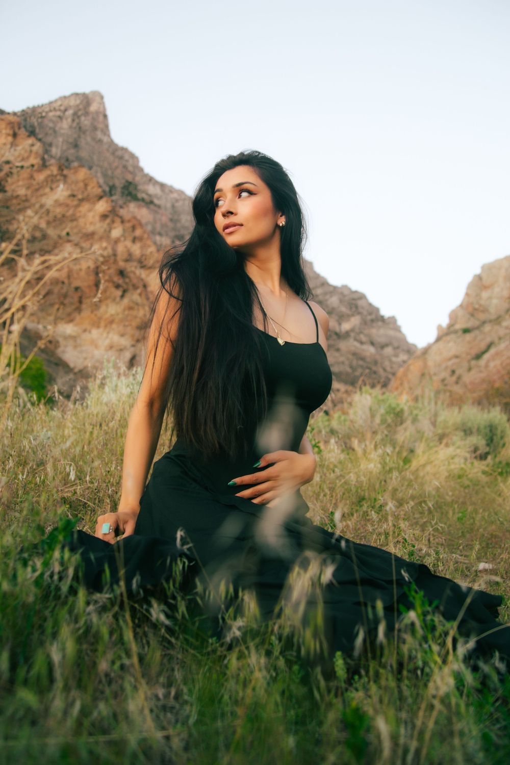 Brunette woman in black sundress sits in grass gazing up at rocky mountain landscape.