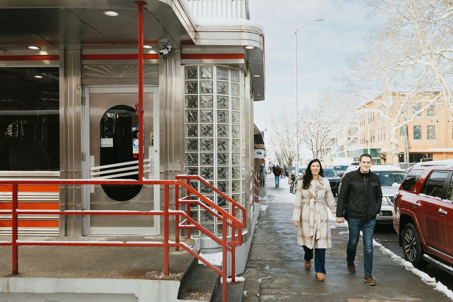 Couple walking together on a snowy urban sidewalk past a modern diner with red railings and glass block windows in winter.