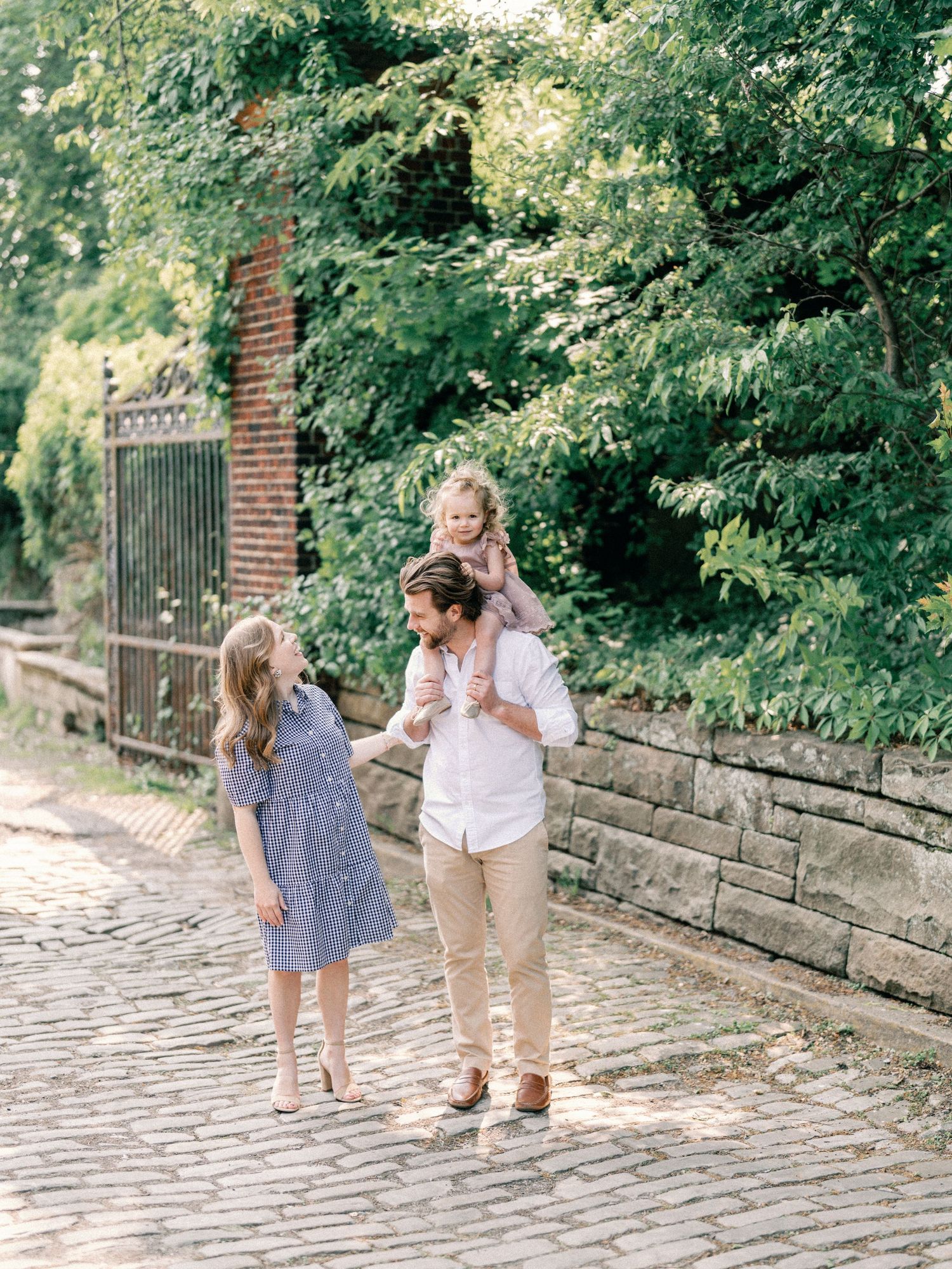 A family of three walks on cobblestone path, dad carrying toddler on shoulders, mom in blue dress, lush greenery behind.