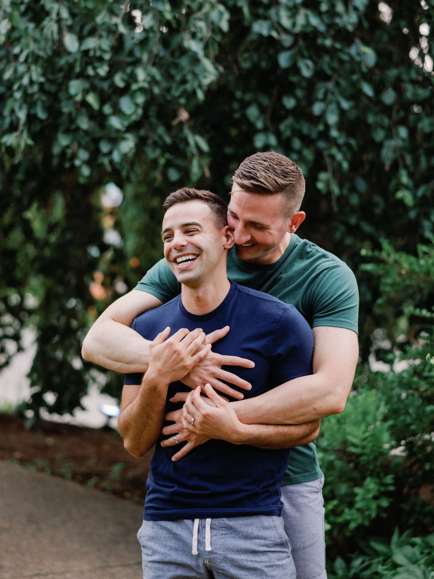 Two men hugging and laughing outdoors, surrounded by lush green trees in a park setting.