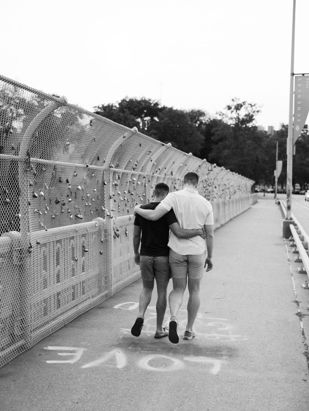 Two men walking arm in arm on a bridge path with love locks on fence, black and white photo.