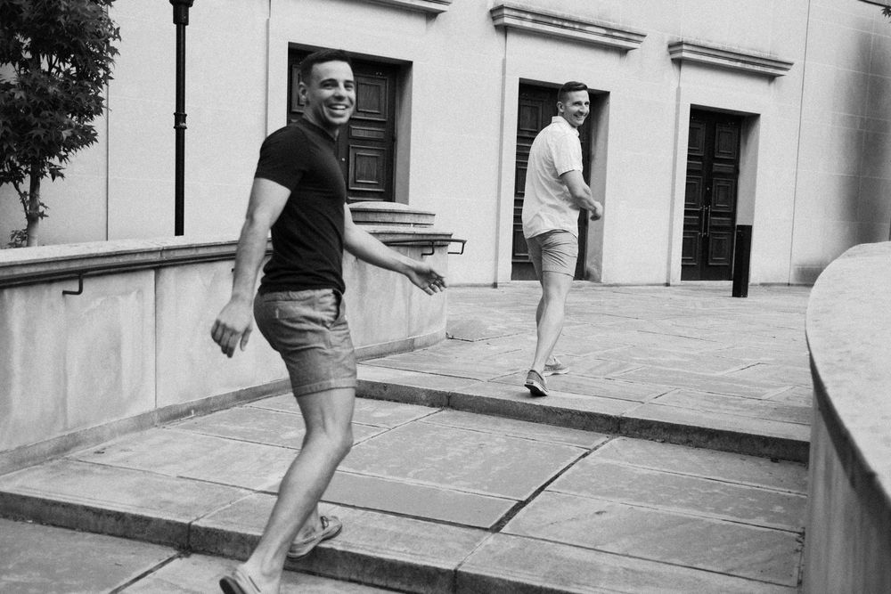 Two men in casual summer outfits walking and smiling on stone steps outside a classic building.
