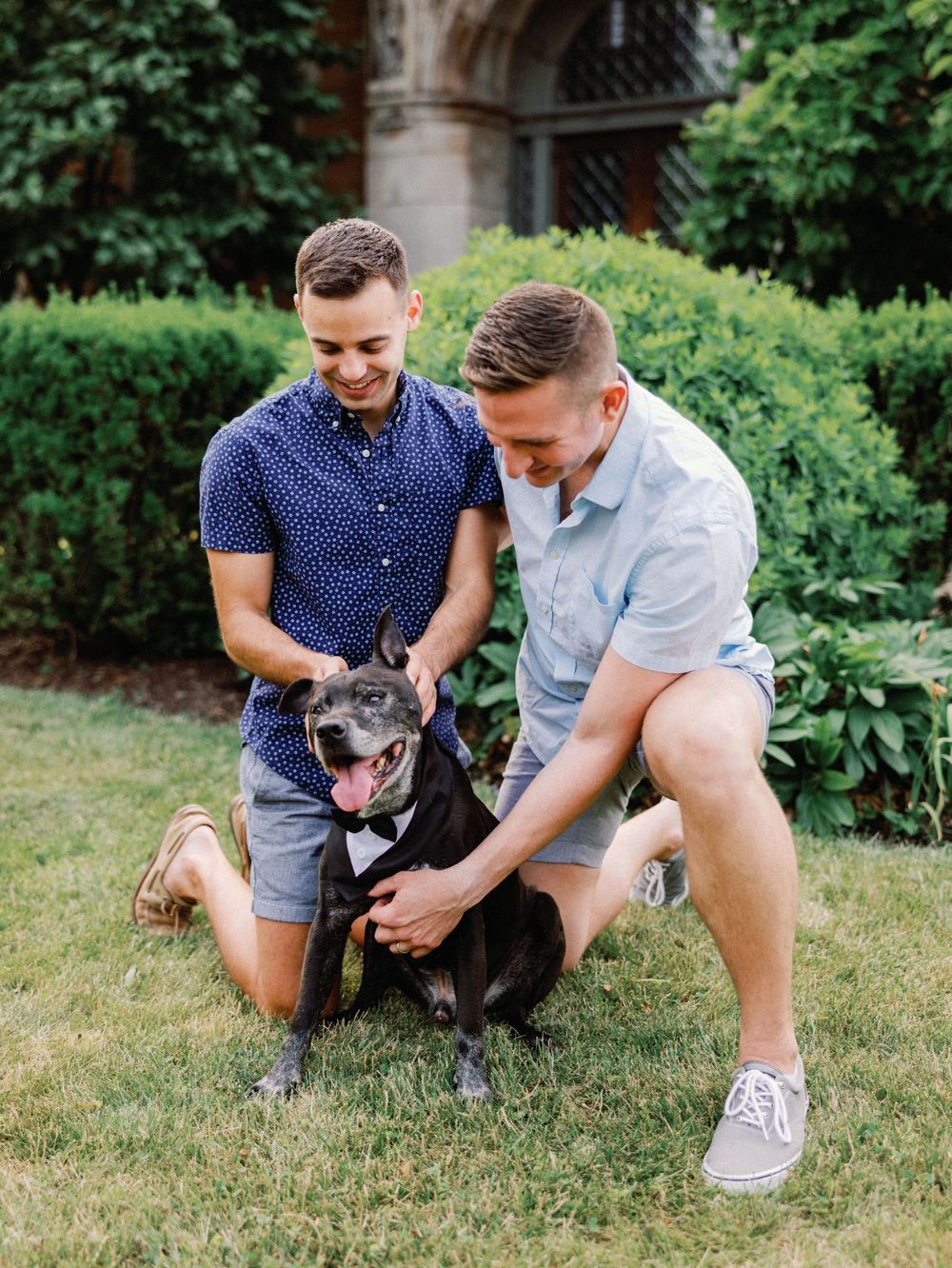 Two men kneel on grass petting a black dog wearing a tuxedo bow tie outdoors near green shrubs.
