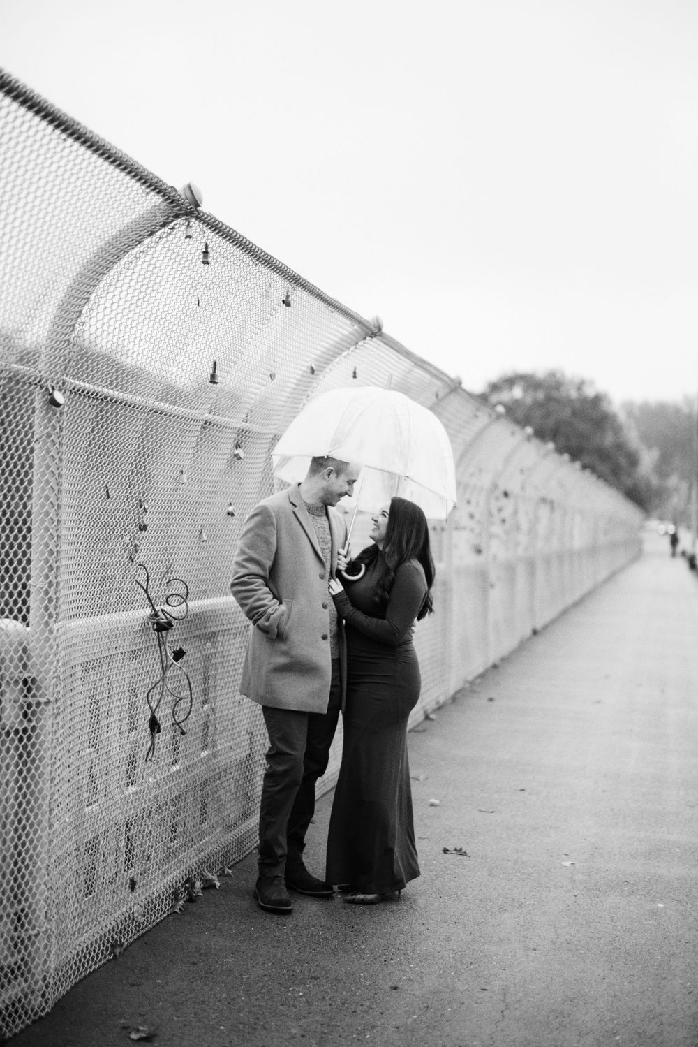 Black and white photo of a couple sharing a clear umbrella on a bridge walkway, gazing at each other romantically.