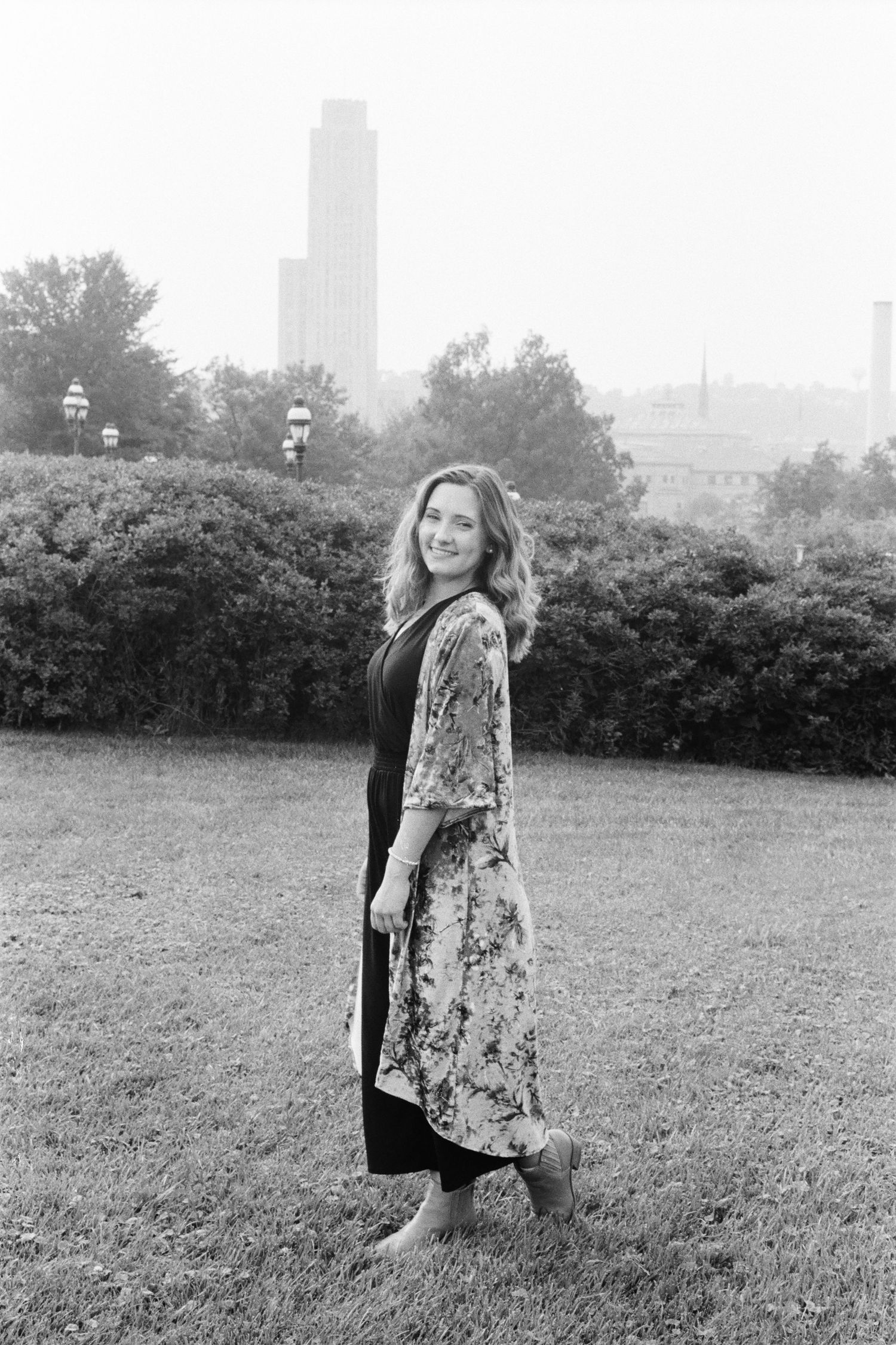 Woman in floral kimono smiling in park with city skyline, black and white photo.