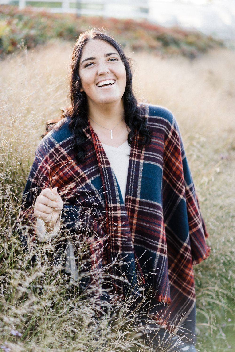 Smiling young woman wrapped in plaid blanket scarf standing in tall golden grass field outdoors.