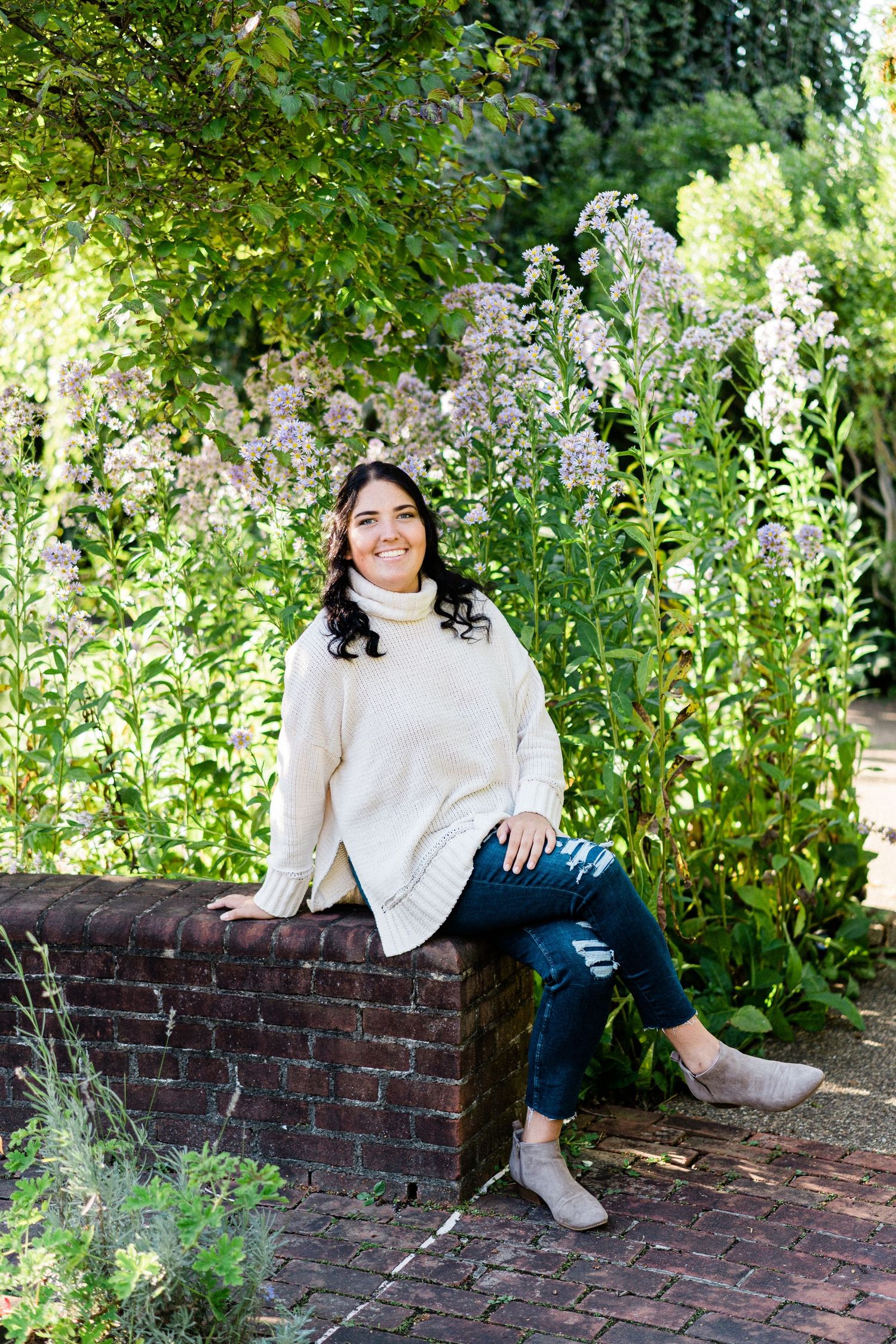 Woman in cream sweater and ripped jeans sitting on brick wall surrounded by lush garden flowers, smiling outdoors.
